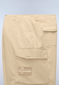 Pantalones cargo beige con bolsillos con solapa y un parche rectangular cosido con un diseño de cruz en el bolsillo delantero.