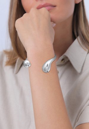 Bracelet en argent au design élégant et fluide. Présente une forme courbée avec deux accents arrondis, une finition métallique polie et une texture lisse.