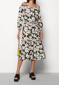 Blumenkleid mit schulterfreier Ausschnitt, versehen mit weißen Lilien auf schwarzem Hintergrund, elastischem Taillenbund und gestuftem Rock. Schwarze Plateaustiefel.