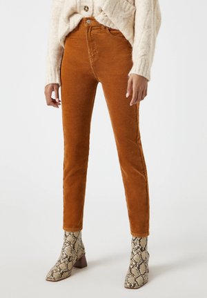 Femme portant un pantalon en velours côtelé taille haute couleur rouille, associé à un pull torsadé crème et des bottines à motif peau de serpent.