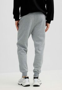 Persona que lleva pantalones deportivos gris claro y zapatillas blancas con detalles en negro, de pie frente a un fondo claro y liso.