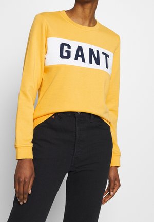 Personne portant une chemise jaune à manches longues avec "GANT" en lettres noires sur une bande blanche, associée à un pantalon noir taille haute.