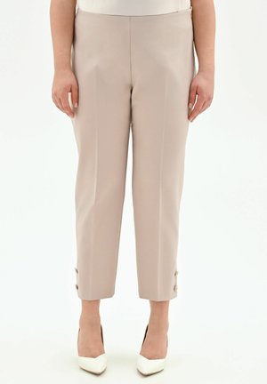 Pantaloni beige su misura alla caviglia con dettagli a bottoni sui polsini, indossati con décolleté bianchi a punta e una maglia bianca.