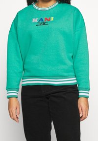 Persona che indossa una felpa turchese con il testo multicolore "KANI" e pantaloni neri, in piedi su uno sfondo bianco.