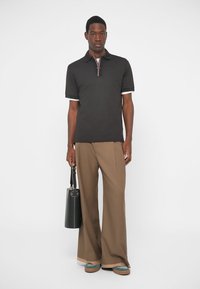 Chemise polo noire avec des accents blancs et un col rayé, associée à un pantalon large marron et des baskets vertes. Tient un sac noir.