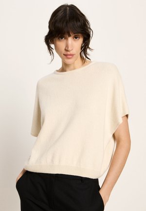 Woman with short dark hair wearing a loose cream short-sleeve sweater and black pants, standing with hands in pockets against plain background.