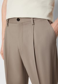 Gros plan sur un pantalon taupe sur mesure avec plis, fermeture par boutons et main dans la poche latérale, porté avec un haut beige rentré.