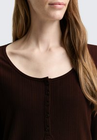 Bruine geribbelde top met een ronde halslijn, zeven knopen en een gladde textuur. Gestyled met lang, golvend haar over de schouder.