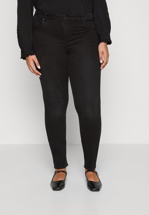 Vrouw draagt strakke zwarte jeans, zwart langarmshirt en zwarte Mary Jane schoenen, staand tegen een effen lichte achtergrond.