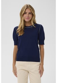 Šviesiaplaukė moteris su laisvais garbanomis, vilkinti tamsiai mėlyną trumpą švarką su baltais siūlų akcentais ir šviesiai rudos spalvos aukštomis kelnėmis, stovinti.