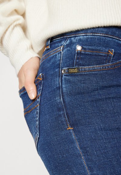 Jeans de mezclilla azul oscuro con costuras naranjas, un aspecto ligeramente desgastado y un cierre de botón plateado con una etiqueta que dice "Tiger of Sweden."