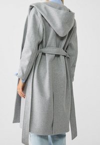 Mujer con un abrigo de lana gris largo y con capucha, con un cinturón atado en la parte de atrás, sobre unos jeans de color azul claro y una camisa de rayas azules.