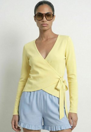 Femme portant de grandes lunettes de soleil marron, un pull cache-cœur jaune noué sur le côté et un short bleu clair à volants, debout devant un fond uni.