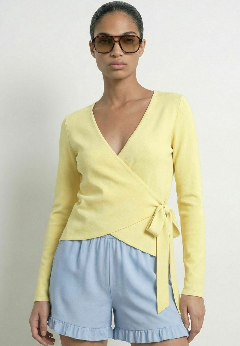 Femme portant de grandes lunettes de soleil marron, un pull cache-cœur jaune noué sur le côté et un short bleu clair à volants, debout devant un fond uni.