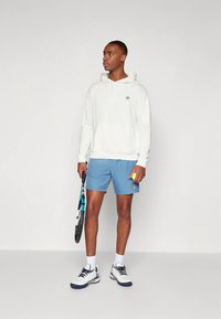 Fehér kapucnis pulóver, kék sportnadrág, teniszütőt és labdát tart a kezében. Cipője fehér, tengerészkék részletekkel. Egyszerű, sportos dizájn.