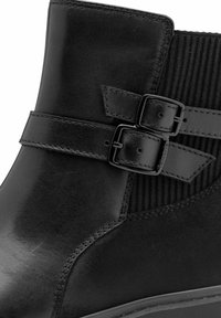 Bottines noires à cheville en cuir lisse, avec des panneaux latéraux en tissu et deux boucles décoratives pour le détail. Semelle en caoutchouc à profil bas.