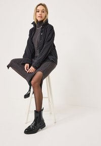 Giacca di denim nera sopra un vestito a maglia grigia con trecce. La modella indossa stivali neri lucidi alla caviglia e collant neri, seduta su uno sgabello bianco.