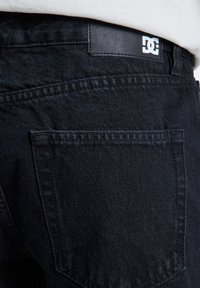 Les jeans en denim noir présentent un tissu texturé, des coutures sur la poche arrière et une étiquette en cuir rectangulaire avec un logo blanc sur la ceinture.