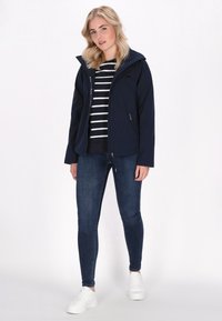 Mladá žena stojí, oblečená v námorníckej bunde cez prúžkované tričko, doplnená tmavými džínsami a bielymi teniskami, na neutrálnej background.