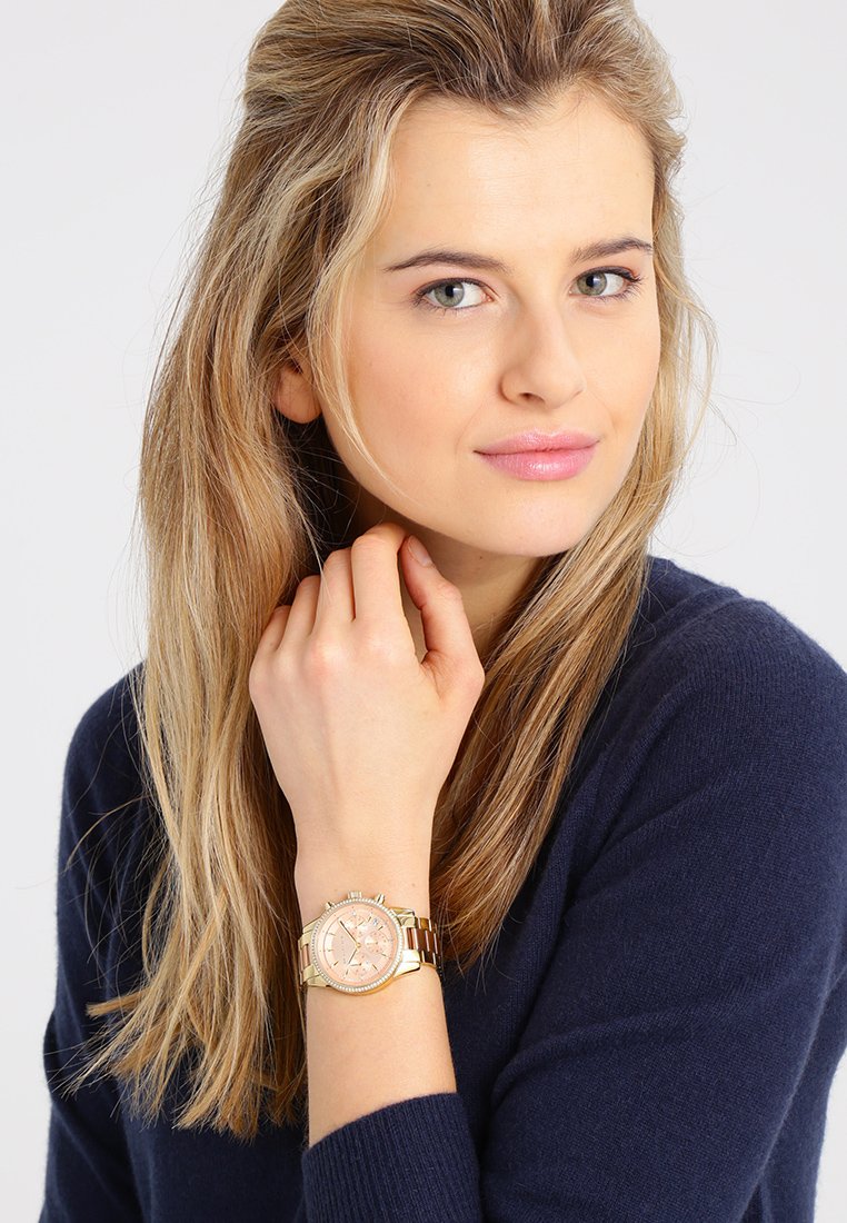 Reloj dorado con una esfera rosa, características de cronógrafo y pulsera bicolor. Lleva puesto en la muñeca, combinado con un suéter oscuro.