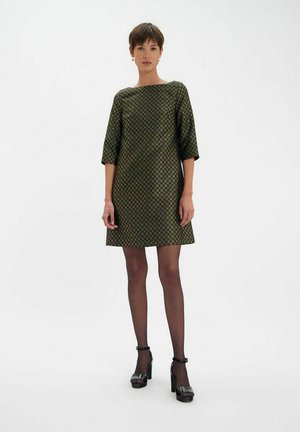 Robe verte à motifs avec des manches trois-quarts, coupe évasée, tissu texturé et motif en losanges discret, associée à des talons noirs et des collants.