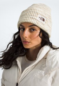 Cremefarbene Strickmütze mit geripptem Muster und einem Logo-Patch; kombiniert mit einer weißen Steppjacke mit Reißverschluss und hohem Kragen.