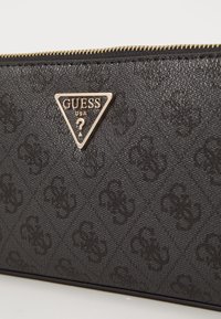 Sort pung med tekstureret overflade, præget logo-mønster og et guldfarvet metaltriangel-logo med et spørgsmålstegn. Lynlåslukning.