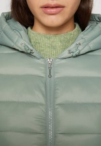 Lysegrøn dunjakke med hætte, der har en frontlynlås og sølvfarvet hardware, lagdelt over en tekstureret grøn sweater.