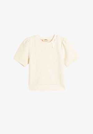 Top corto a maniche corte di colore crema con tessuto testurizzato, scollatura rotonda e un piccolo nastro bianco decorativo sulla spalla sinistra.