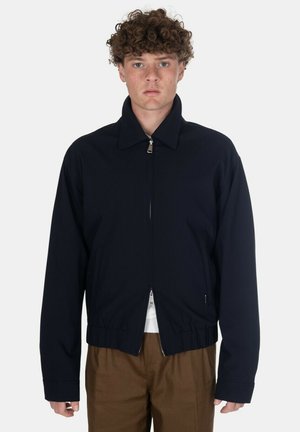 Giovane uomo con capelli ricci che indossa una giacca blu scuro con zip e pantaloni marroni, in piedi davanti a uno sfondo bianco semplice.