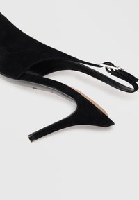 Czarny zamszowy but na wysokim obcasie typu slingback z ostro zakończonym noskiem i regulowanym paskiem na klamrę na białym tle.