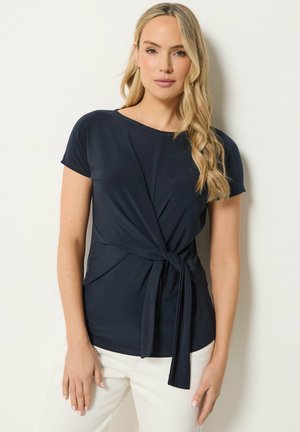 Femme aux longs cheveux blonds portant un chemisier bleu marine à manches courtes noué à la taille et un pantalon blanc, debout devant un fond clair uni.