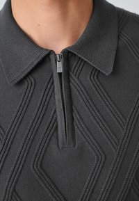 Sötét szürke pulóver, gallérral, félig cipzárral és texturált geometrikus mintával a mellkason. Kötött anyagból készült, bordázott részletekkel.