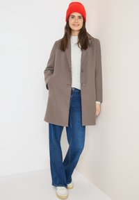 Cappotto di lana marrone con un design sartoriale, due bottoni, tasche laterali, abbinato a un maglione grigio chiaro e jeans a zampa d'elefante blu. Berretto di lana rosso.