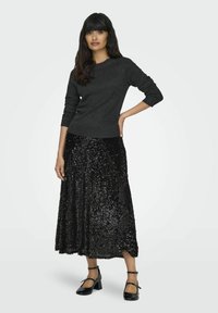 Maglione di carbone con maniche lunghe abbinato a una gonna maxi nera con paillettes. La modella indossa tacchi a spillo neri in vernice con cinturini alla caviglia.