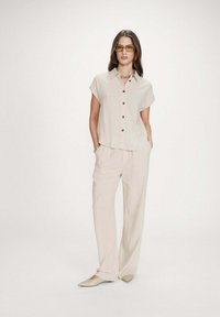 Mujer con camisa beige de manga corta abotonada, pantalones anchos a juego, zapatos puntiagudos, gafas de sol con lentes amarillas y joyas doradas.