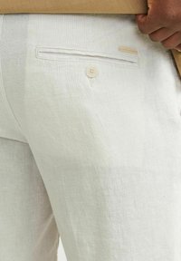 Un pantalon en lin texturé blanc doté d'une poche arrière avec fermeture à bouton et d'un accent de marque. Le tissu présente un léger motif côtelé.