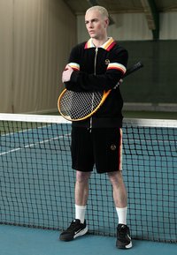 Mustast veluurist treeningkomplektist mitmevärviliste triipudega, mustad lühikesed püksid ja valged sportesocksid. Hoides tennisereketit, seisab võrgu kõrval.
