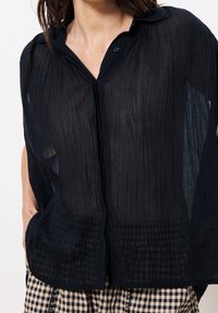 Femme portant un chemisier transparent noir à boutons avec des lignes verticales texturées et un pantalon beige et noir à carreaux, mains dans les poches.