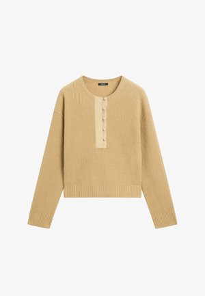 Strikket sweater i lys beige med lange ærmer og ribbet struktur. Har en knappet slids i et kontrastfarvet stof foran.