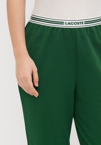 Grønne bomullsjoggesko med et hvitt og grønt stripete elastisk midjebånd merket "LACOSTE." Har en avslappet passform og en glatt tekstur.
