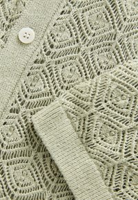 Stickad kofta i ljusgrönt med ett hexagonalt spetsmönster, utrustad med en texturerad stickad krage och en rund vit knappstängning.