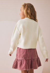 Balts īss džemperis ar ribveida aprocēm, kombinēts ar rozā korduroja svārkiem ar volānu apakšmalā, skatīts no mugurpuses.