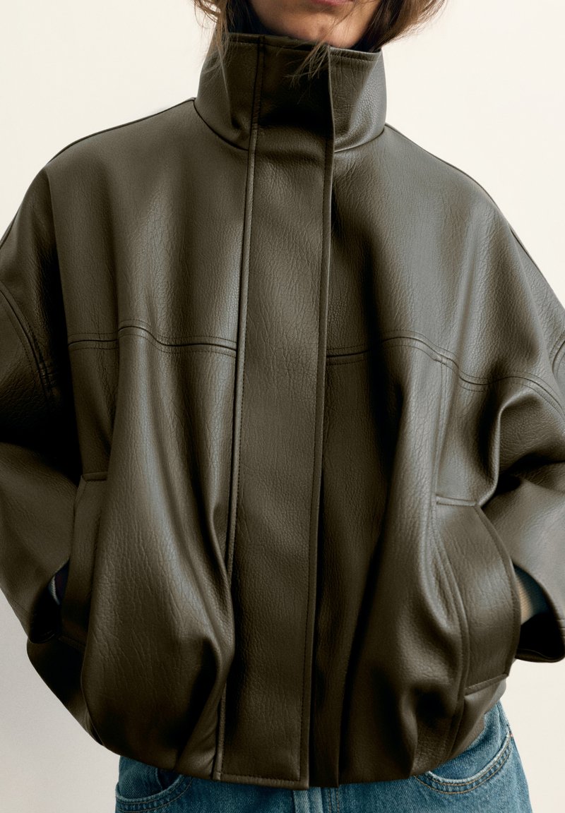 Personne portant une veste en cuir vert olive foncé surdimensionnée avec un col montant et les mains dans les poches, associée à un jean bleu.