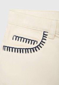 Krämfärgad bomullstyg med marinblå broderade sömmar längs fickkanterna. Designen har en klassisk fickform och textur.