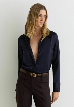 Femme aux cheveux blonds ondulés mi-longs, portant une chemise en soie bleu marine déboutonnée en haut et un pantalon marron foncé avec une ceinture.