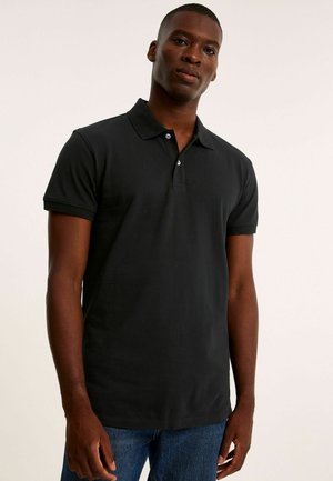 Mann trägt ein schlichtes schwarzes Polohemd mit kurzen Ärmeln und blaue Jeans, steht vor einem schlichten hellen Hintergrund.