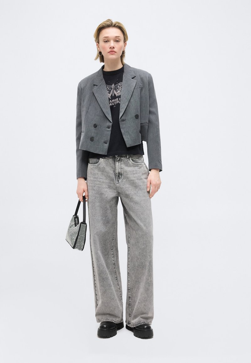 Jeune personne portant un blazer gris court, un t-shirt graphique noir, un jean gris ample, des chaussures à plateforme noires, tenant un petit sac à main.