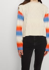 Personne portant un pantalon noir, un pull sans manches crème en maille torsadée par-dessus une chemise à manches longues avec des rayures horizontales orange, blanches et bleues.