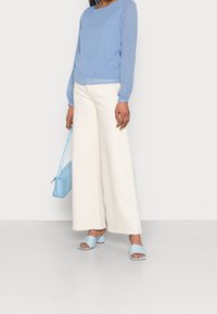 Blusa azzurra chiara con texture e maniche lunghe, abbinata a pantaloni larghi di un bianco sporco. Gli accessori includono una borsa azzurra chiara e sandali coordinati.
